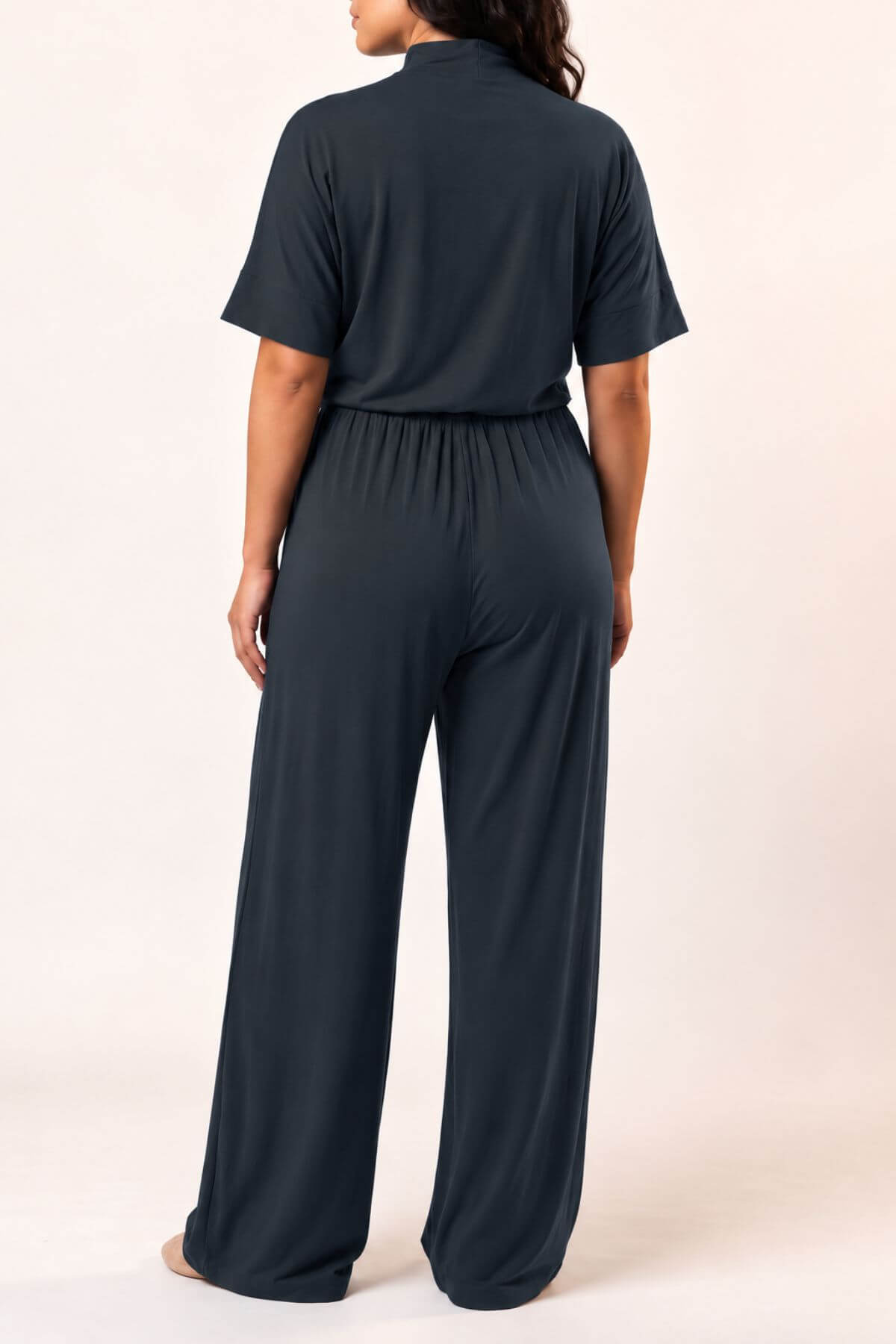 A woman viewed from the back wearing a dark charcoal gray short-sleeve jumpsuit with a high mock neck and wide-leg pants, standing against a plain light beige studio background.