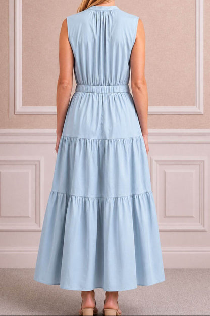A woman viewed from the back wearing a sleeveless light blue tiered maxi dress with a gathered waist, standing in front of a beige paneled wall.
