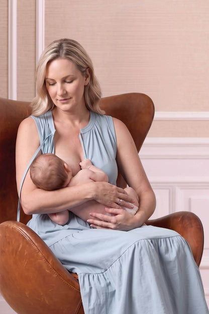 A woman in a light blue sleeveless dress sits in a brown leather armchair, breastfeeding a baby wrapped in a white blanket. The background features a soft beige paneled wall.