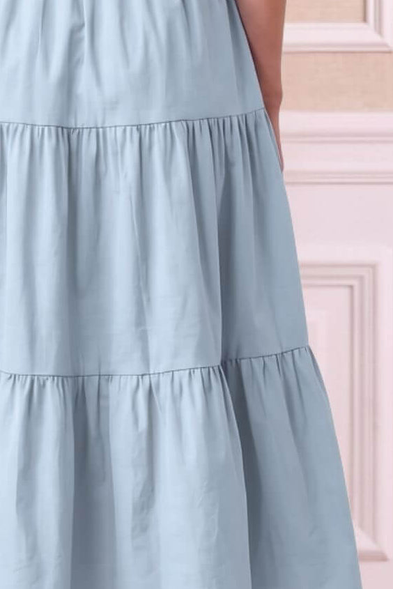 A woman shown from the back wearing a sleeveless light blue tiered maxi dress with a gathered waist, set against a soft beige paneled wall.