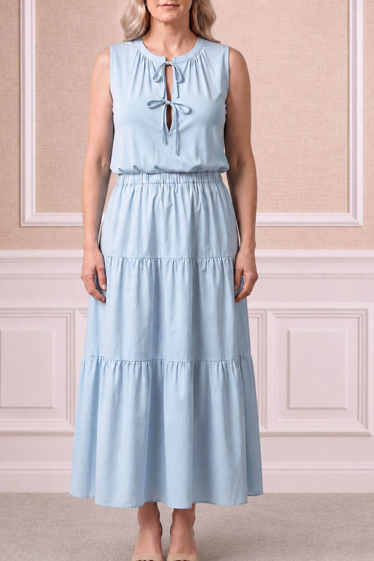 A woman wearing a sleeveless light blue maxi dress with a tiered skirt and a tie-neck detail, standing in front of a beige paneled wall.