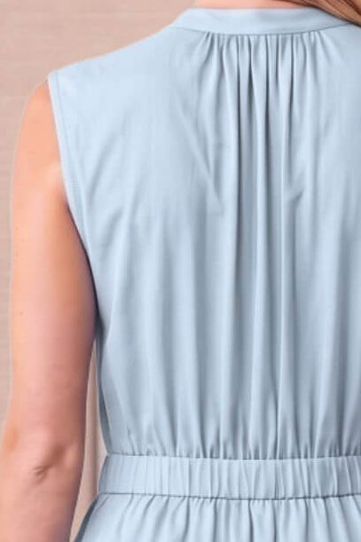 A woman shown from the back wearing a sleeveless light blue tiered maxi dress with a gathered waist, set against a soft beige paneled wall.