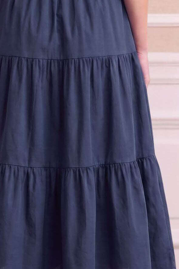 A sleeveless navy blue maxi dress with a tiered skirt and gathered waist, shown from the back on a model against a beige paneled wall.