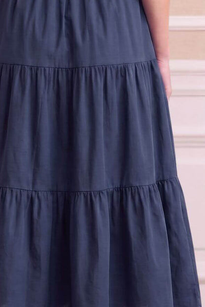 A sleeveless navy blue maxi dress with a tiered skirt and gathered waist, shown from the back on a model against a beige paneled wall.