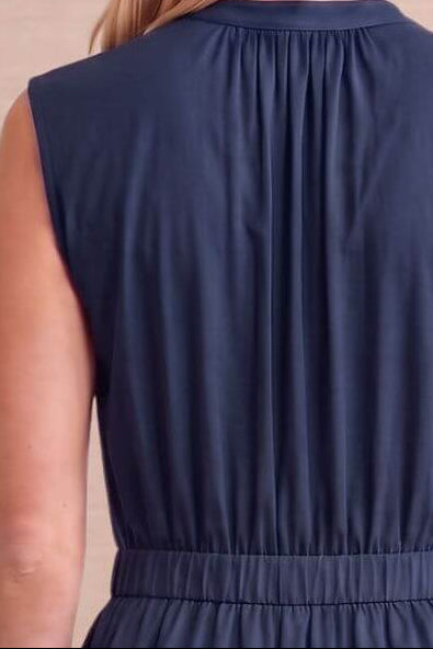 A sleeveless navy blue maxi dress with a tiered skirt and gathered waist, shown from the back on a model against a beige paneled wall.