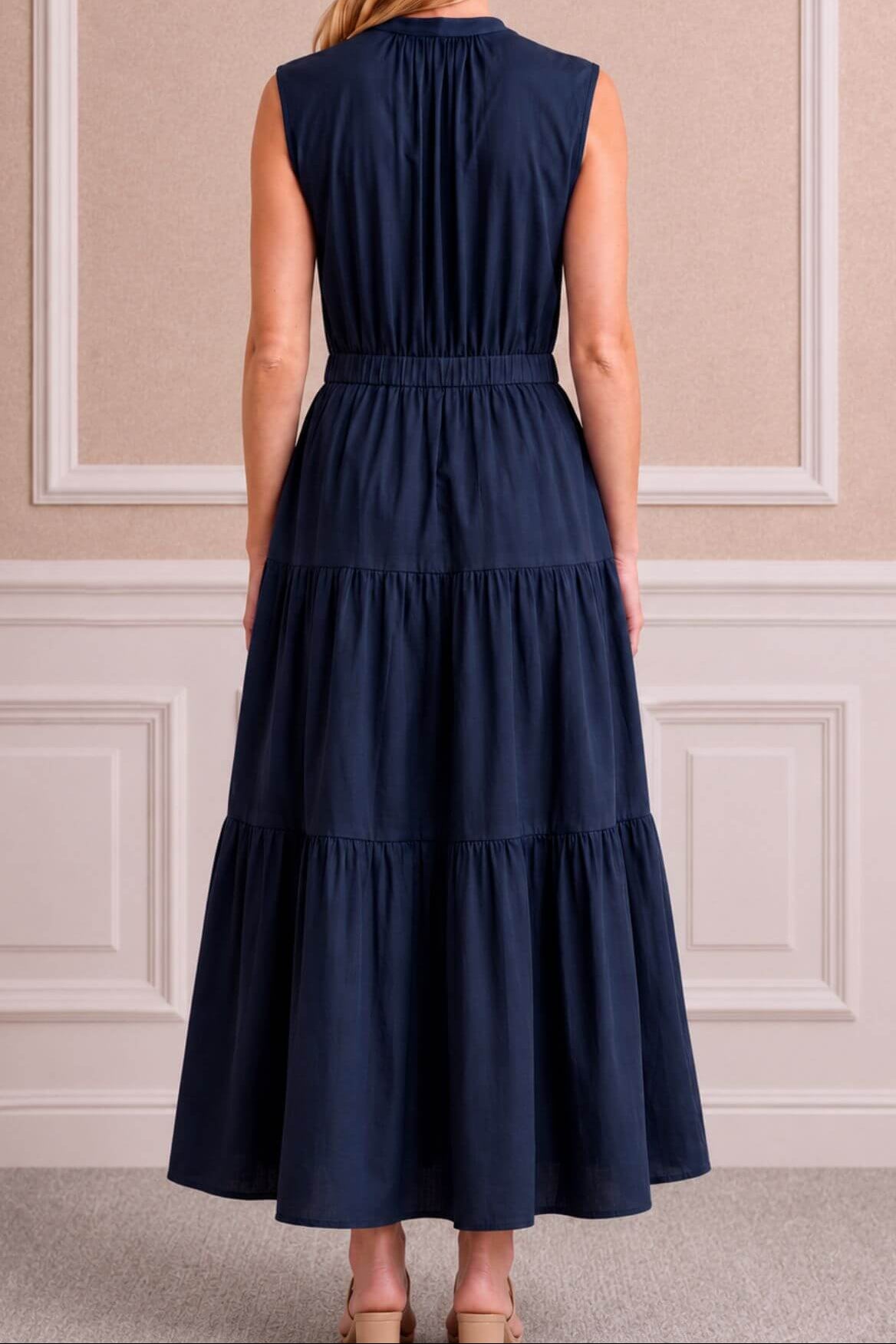 A woman viewed from the back wearing a sleeveless navy blue maxi dress with a gathered waist and tiered skirt, standing in front of a beige paneled wall.