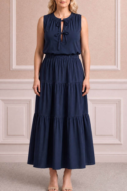 A woman modeling a sleeveless navy blue maxi dress with a tiered skirt and a keyhole tie-neck detail, standing against a beige paneled wall.