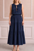 A woman modeling a sleeveless navy blue maxi dress with a tiered skirt and a keyhole tie-neck detail, standing against a beige paneled wall.