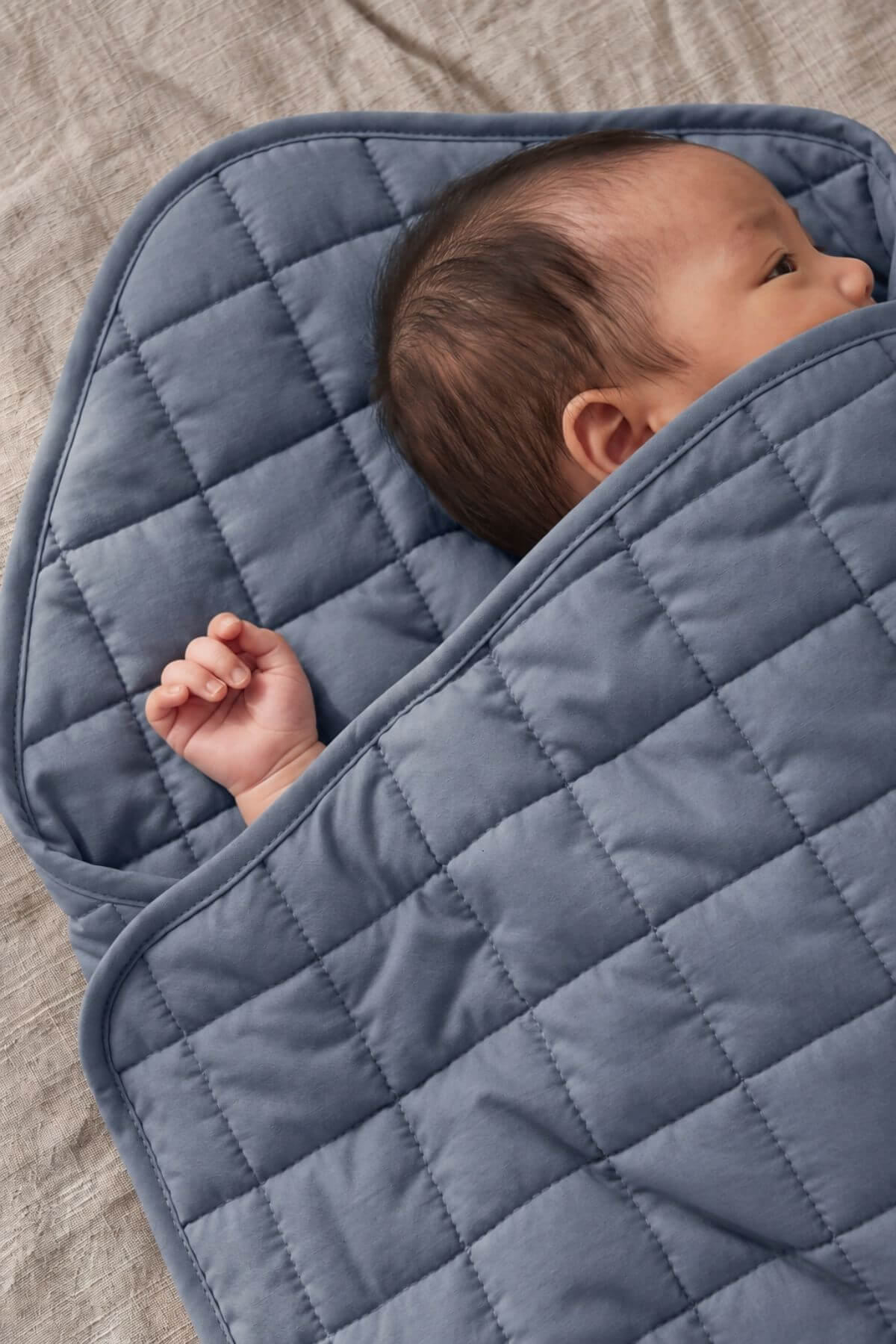 A baby wrapped in a dusty blue quilted blanket, lying on a textured beige fabric surface.