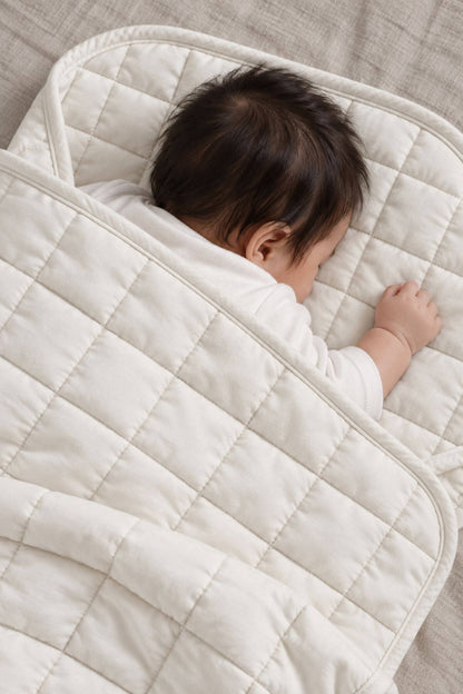 A baby wrapped in a cream-colored quilted blanket, lying on a textured beige fabric surface.