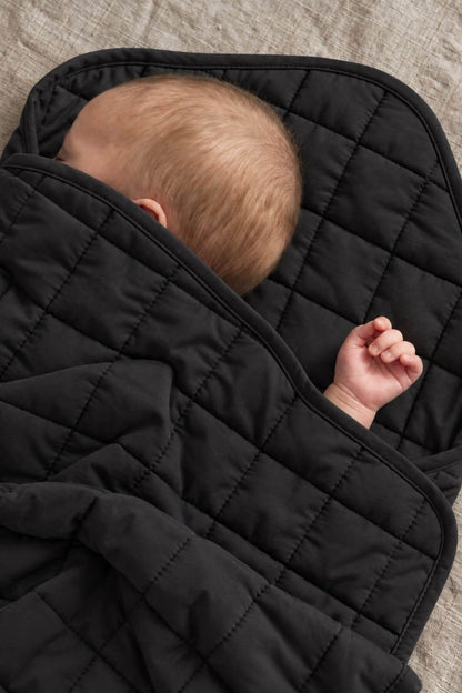 A close-up of a baby wrapped in a black quilted blanket, lying on a textured beige fabric surface.