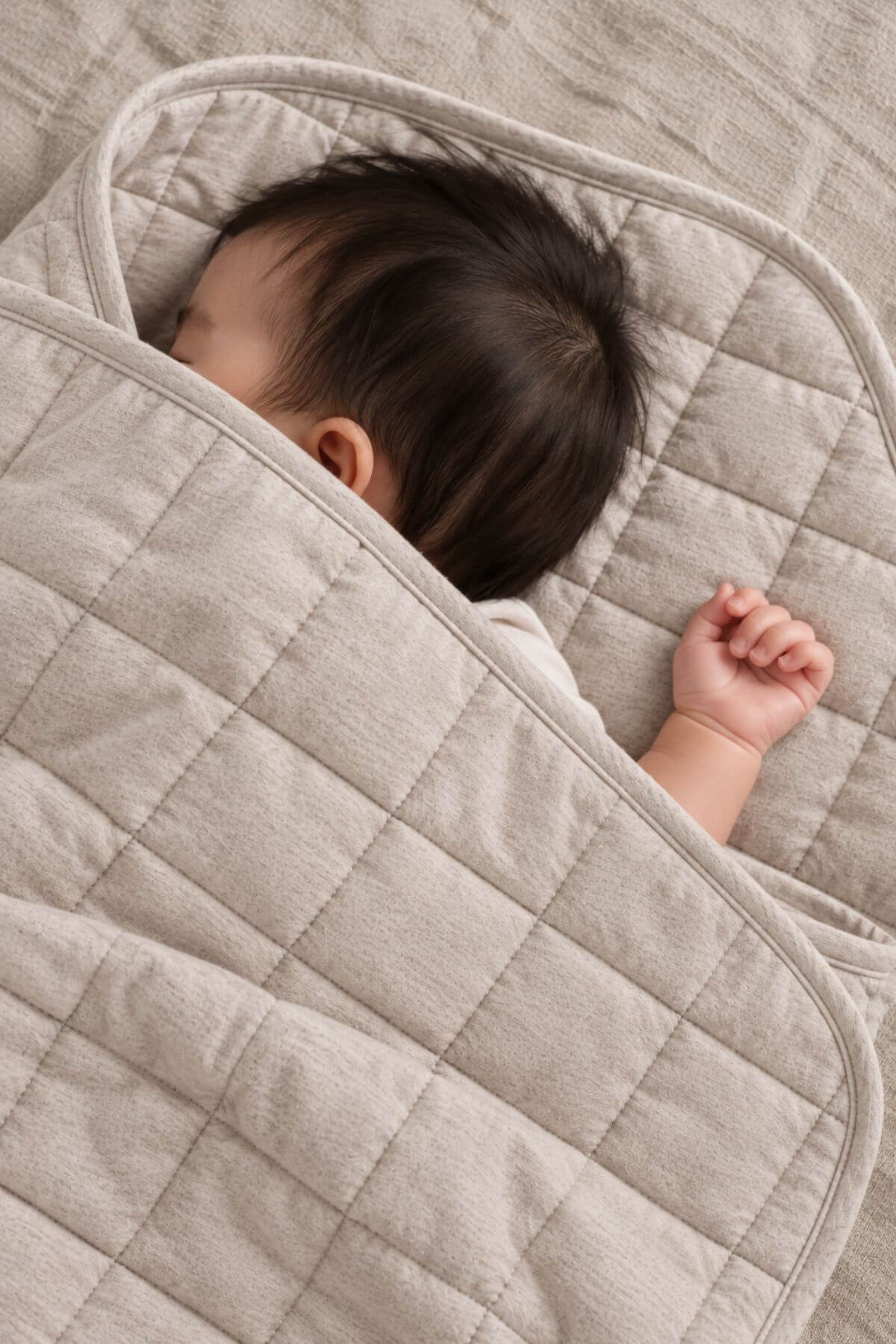 A close-up of a baby wrapped in a quilted beige blanket, lying on a matching textured beige bedding surface.