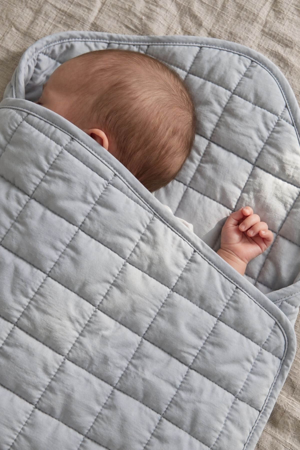A baby swaddled in a light gray quilted blanket, lying on a textured beige linen surface.