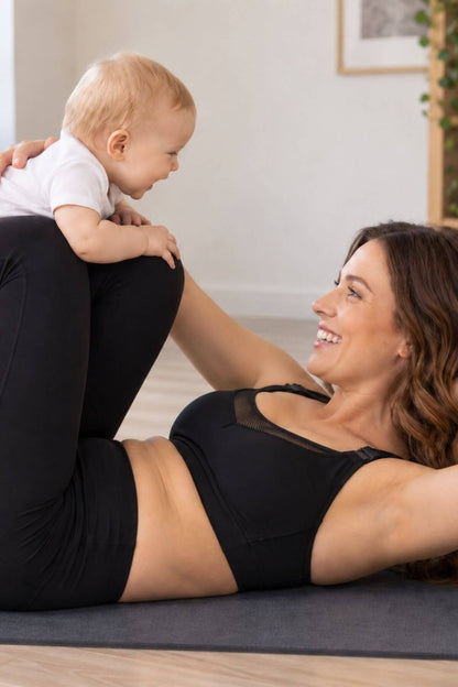 A woman in the supermom sweetheart mesh racerback nursing &amp; hands-free pumping sports bra and leggings lies on a yoga mat while holding a baby who is wearing a white t-shirt and black pants. They are smiling and interacting in a bright, modern indoor setting.