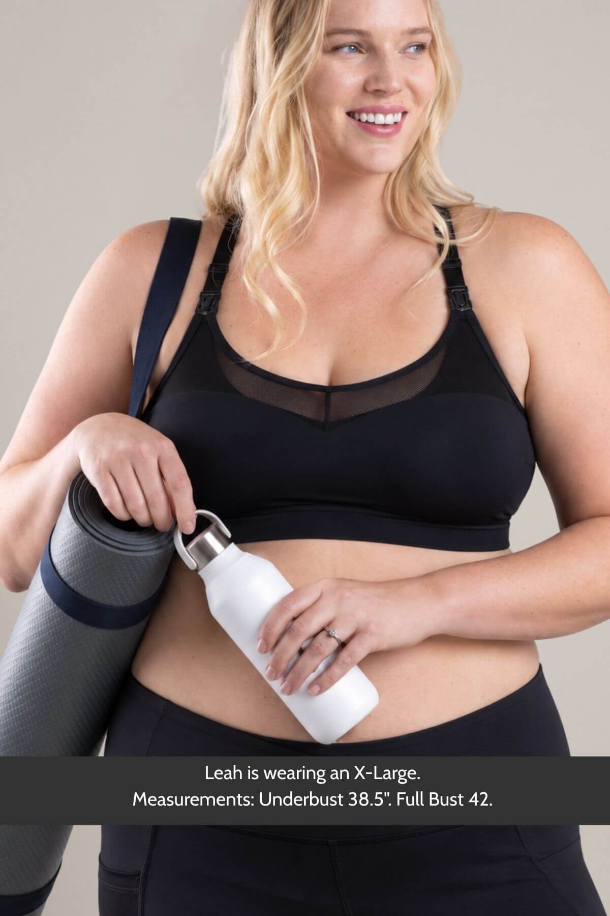 A smiling blonde woman wearing the supermom sweetheart mesh racerback nursing &amp; hands-free pumping sports braand black bottoms, holding a rolled gray exercise mat and a white water bottle. Text at the bottom reads: &
